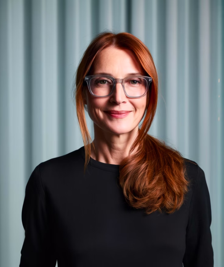Smiling woman with red hair tied in a side ponytail, wearing glasses and a black top, standing against a light blue curtain background.