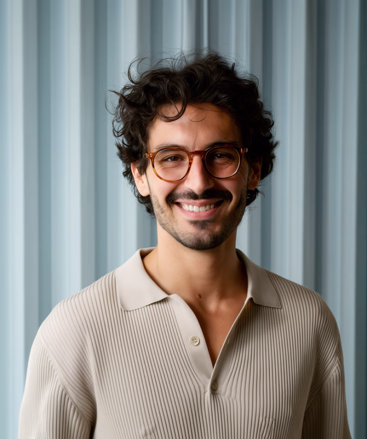 Smiling man with curly hair and glasses wearing a beige ribbed shirt in front of a blue-gray corrugated background.